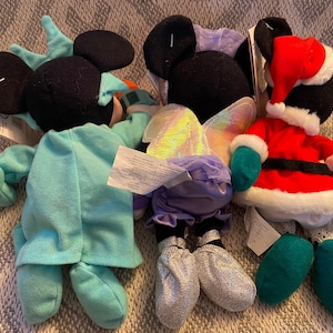 May include: Three plush Mickey Mouse toys dressed in different costumes. One is wearing a blue and white Statue of Liberty costume, another is wearing a purple fairy costume with iridescent wings, and the third is wearing a red and green Santa Claus costume.