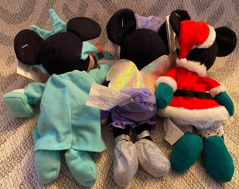 May include: Three plush Mickey Mouse toys dressed in different costumes. One is wearing a blue and white Statue of Liberty costume, another is wearing a purple fairy costume with iridescent wings, and the third is wearing a red and green Santa Claus costume.