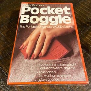 May include: A vintage Parker Brothers Pocket Boggle game box. The box is orange and brown with the game title in large white letters. The image shows a hand holding a red game piece. Text on the box reads: "The Portable 3-Minute WORD Game."