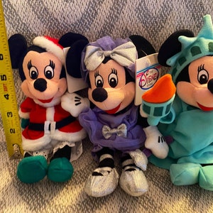 May include: Three plush Mickey Mouse toys dressed in different outfits. The first is wearing a red and white Santa Claus suit with green shoes. The second is wearing a purple dress with a silver bow and silver shoes. The third is wearing a blue and green Statue of Liberty costume.
