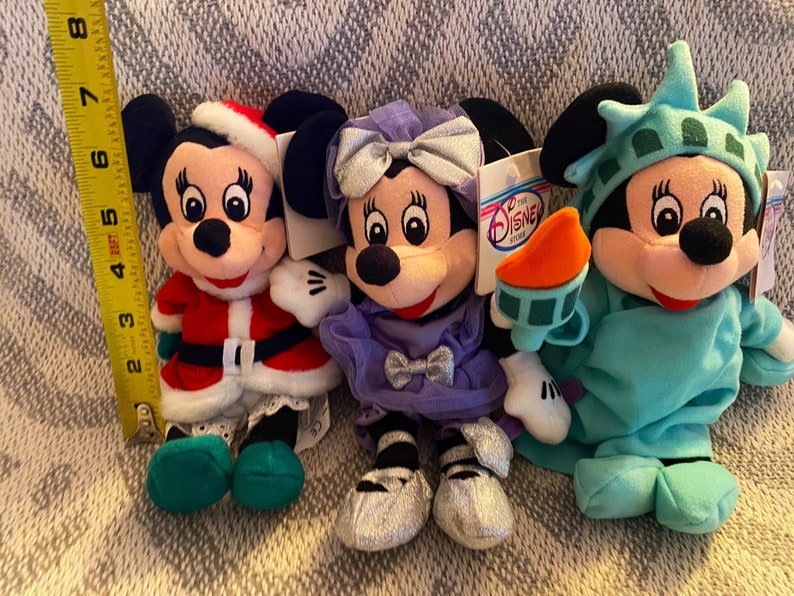 May include: Three plush Mickey Mouse toys dressed in different outfits. The first is wearing a red and white Santa Claus suit with green shoes. The second is wearing a purple dress with a silver bow and silver shoes. The third is wearing a blue and green Statue of Liberty costume.