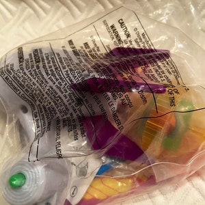 May include: A clear plastic bag containing various colorful toys, including purple, yellow, and green components. The bag has printed text and warnings, including a caution about small parts and suffocation hazards. The toys appear to be from the Disney Pixar Toy Story franchise.