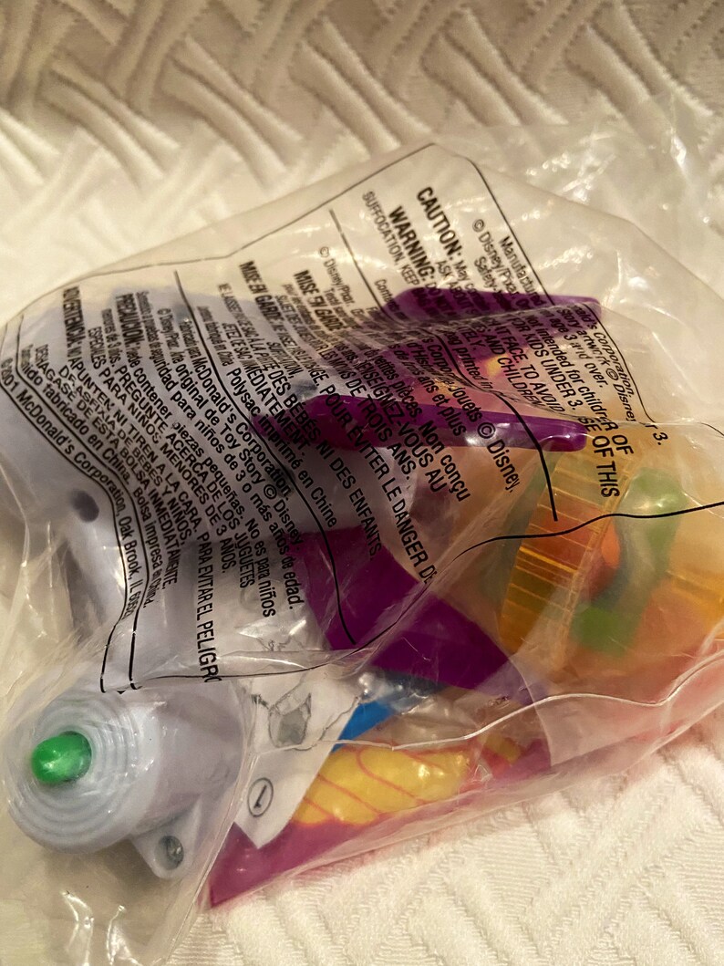 May include: A clear plastic bag containing various colorful toys, including purple, yellow, and green components. The bag has printed text and warnings, including a caution about small parts and suffocation hazards. The toys appear to be from the Disney Pixar Toy Story franchise.