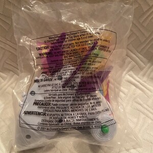 May include: Sealed plastic bag containing a Toy Story collectible toy. The bag has printed warnings and copyright information. The toy is a light purple and green color. The bag is sealed and appears to be from 2001.