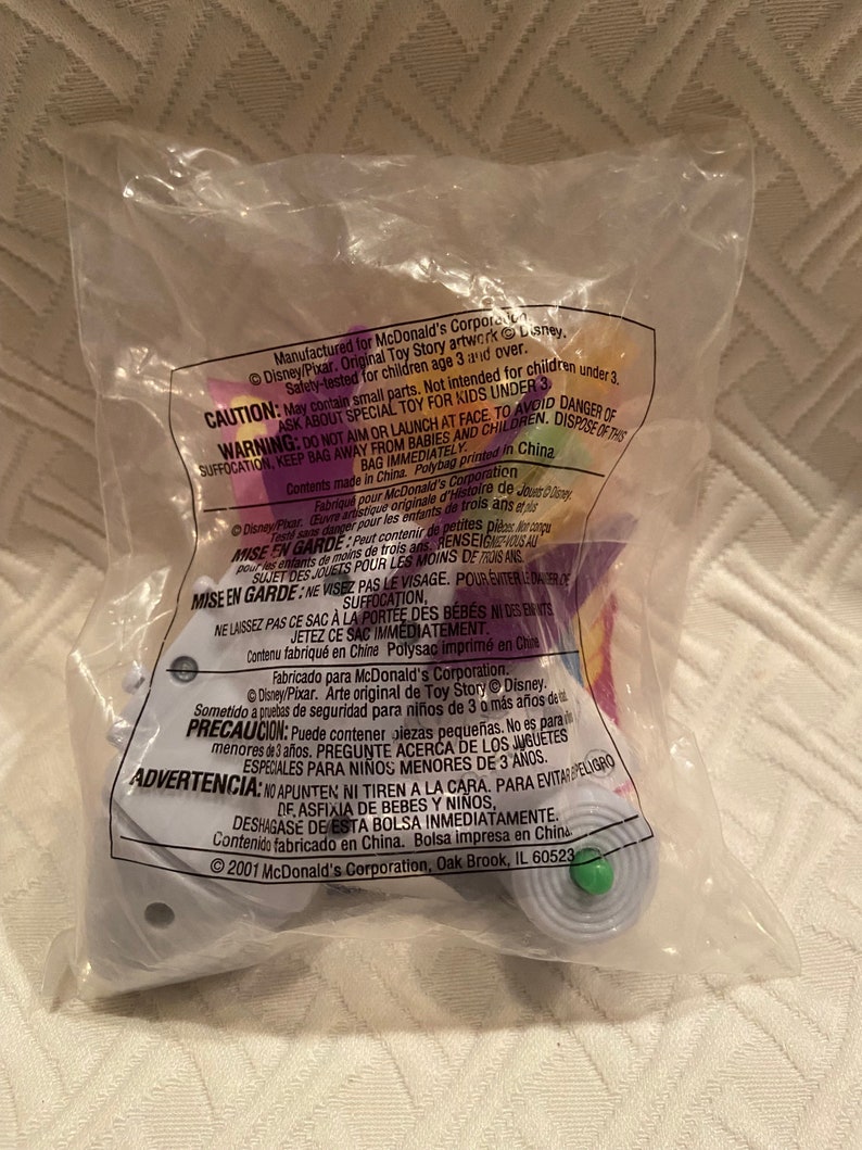 May include: Sealed plastic bag containing a Toy Story collectible toy. The bag has printed warnings and copyright information. The toy is a light purple and green color. The bag is sealed and appears to be from 2001.
