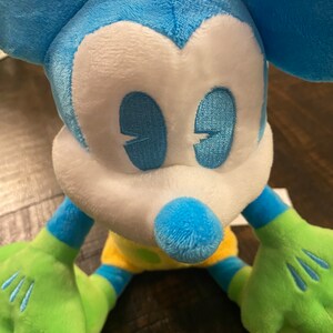 Op de afbeelding: Een blauw en groen pluche Mickey Mouse speelgoed met gele en groene accenten. Het speelgoed heeft een wit gezicht met blauwe ogen en een zwarte neus.