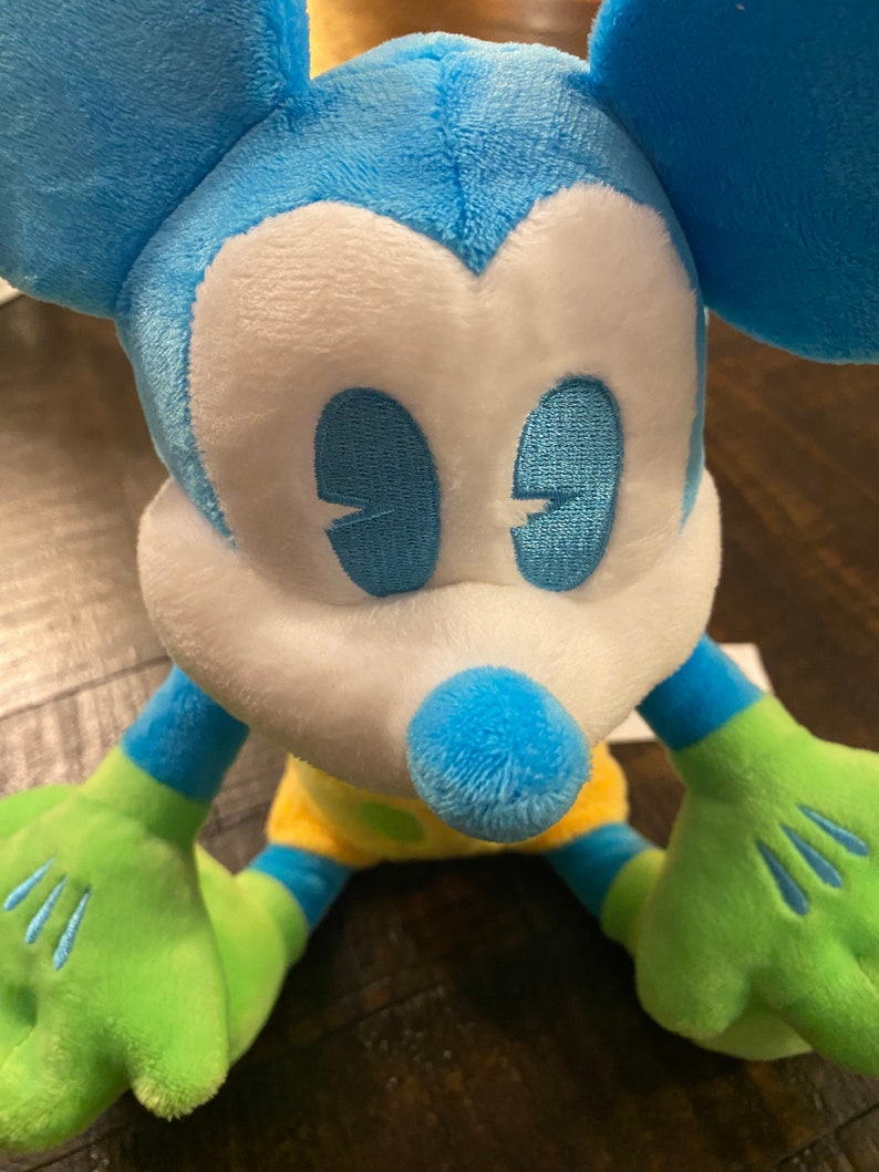 Op de afbeelding: Een blauw en groen pluche Mickey Mouse speelgoed met gele en groene accenten. Het speelgoed heeft een wit gezicht met blauwe ogen en een zwarte neus.