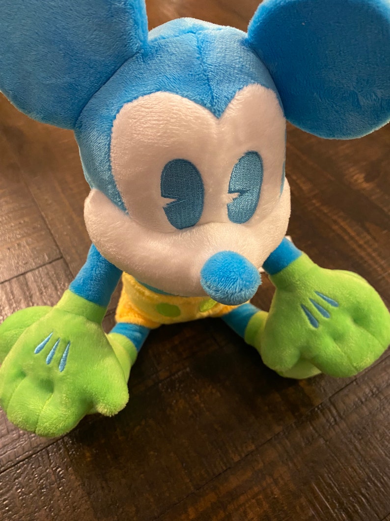 Op de afbeelding: Een blauw en groen pluche Mickey Mouse speelgoed met een gele en groene outfit. Het speelgoed heeft een wit gezicht met blauwe ogen en een zwarte neus.