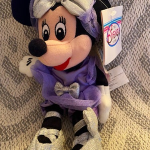 May include: A purple and silver plush Minnie Mouse doll wearing a dress and shoes. The doll has a bow in her hair and a tag that reads "The Disney Store".