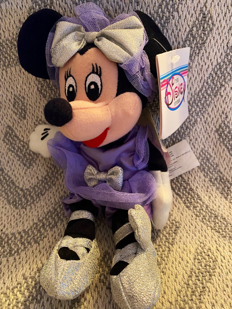 May include: A purple and silver plush Minnie Mouse doll wearing a dress and shoes. The doll has a bow in her hair and a tag that reads "The Disney Store".