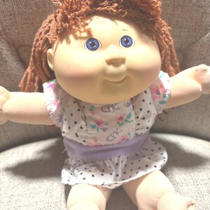May include: A doll with yarn-like auburn hair and blue eyes, wearing a white dress featuring a black polka dot pattern, pink and green floral accents, and the letters CPK printed on the fabric, with a solid light purple waistband.