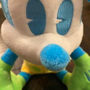Op de afbeelding: Een blauw en groen pluche speelgoed in de vorm van Mickey Mouse. Het speelgoed heeft een wit gezicht met blauwe geborduurde ogen en een blauwe neus.