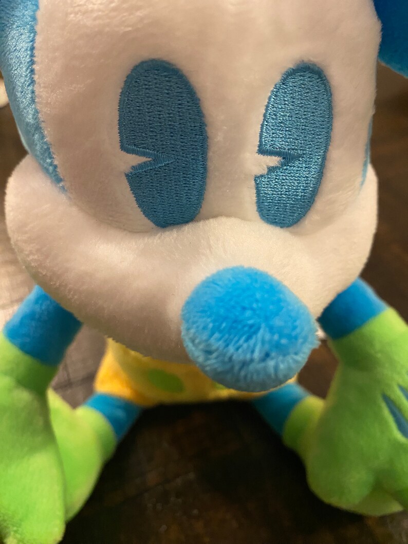 Op de afbeelding: Een blauw en groen pluche speelgoed in de vorm van Mickey Mouse. Het speelgoed heeft een wit gezicht met blauwe geborduurde ogen en een blauwe neus.