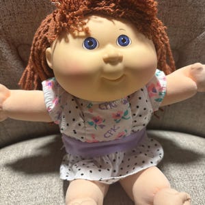 May include: A vintage Cabbage Patch Kid doll with auburn yarn hair and blue eyes. The doll wears a white dress with black polka dots, a purple waistband, and the letters "CPK" printed on the dress.