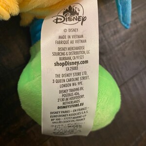 Op de afbeelding: Geel, groen en blauw pluche speelgoed met een Disney-label eraan bevestigd. Het label bevat het Disney-logo, de tekst "Made in Vietnam", "shopDisney.com" en contactgegevens voor Disney-winkels in de Verenigde Staten, het Verenigd Koninkrijk, Nederland en Frankrijk.