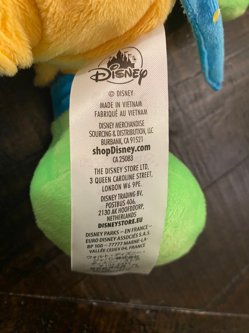 Op de afbeelding: Geel, groen en blauw pluche speelgoed met een Disney-label eraan bevestigd. Het label bevat het Disney-logo, de tekst "Made in Vietnam", "shopDisney.com" en contactgegevens voor Disney-winkels in de Verenigde Staten, het Verenigd Koninkrijk, Nederland en Frankrijk.