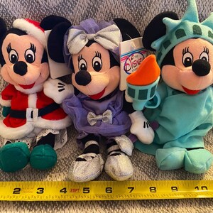 May include: Three plush Mickey Mouse toys dressed in different costumes. The first is wearing a red and white Santa Claus outfit, the second is wearing a purple dress with a silver bow, and the third is wearing a green Statue of Liberty costume.