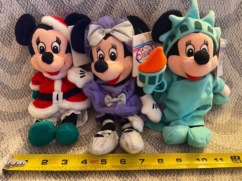 May include: Three plush Mickey Mouse toys dressed in different costumes. The first is wearing a red and white Santa Claus outfit, the second is wearing a purple dress with a silver bow, and the third is wearing a green Statue of Liberty costume.