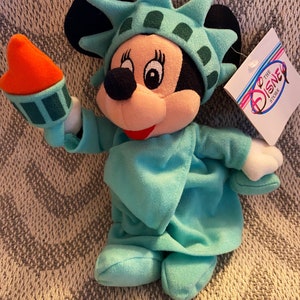 May include: A plush toy of Mickey Mouse dressed as the Statue of Liberty. Mickey is wearing a green dress and a crown with a torch in her hand. The toy has a tag that says "To the Disney Store".