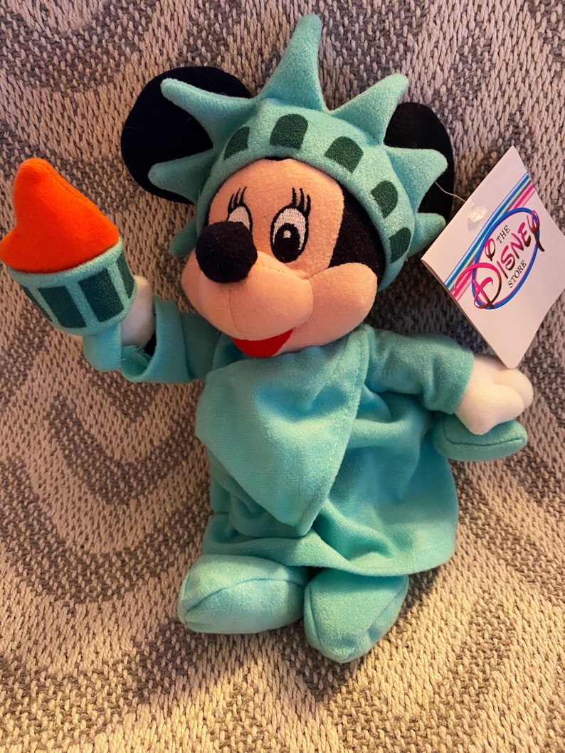 May include: A plush toy of Mickey Mouse dressed as the Statue of Liberty. Mickey is wearing a green dress and a crown with a torch in her hand. The toy has a tag that says "To the Disney Store".