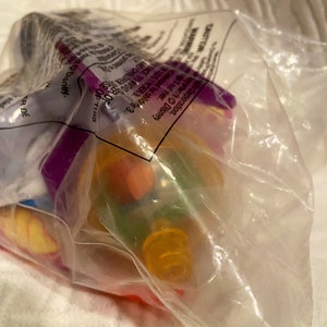 May include: A clear plastic bag containing a colorful toy. The toy appears to have orange, yellow, green, and purple elements. The bag has text that includes the word "Disney".
