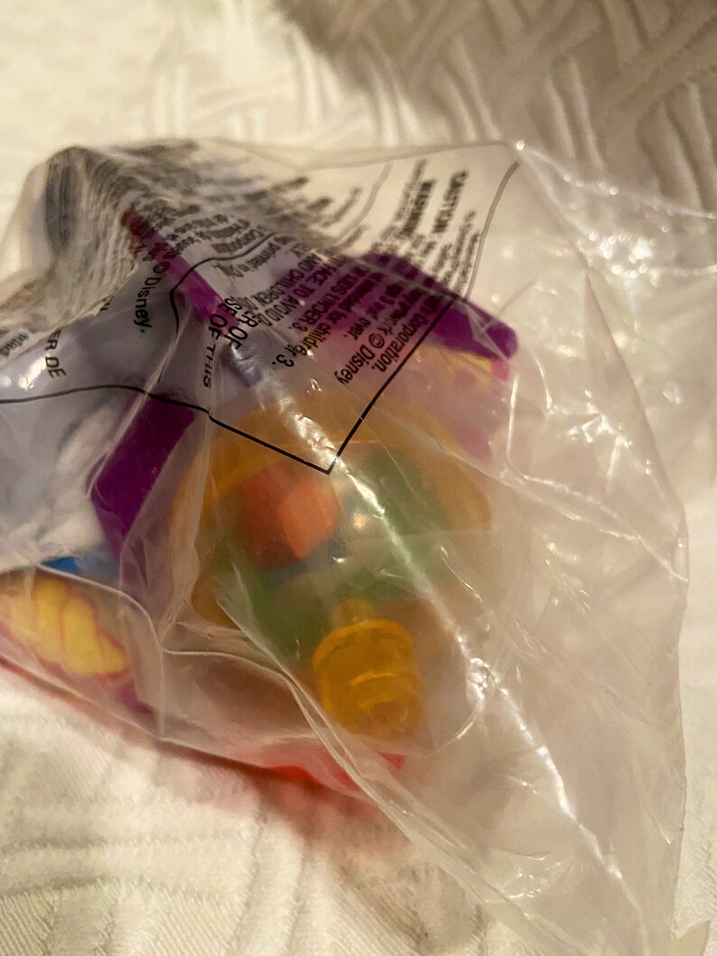 May include: A clear plastic bag containing a colorful toy. The toy appears to have orange, yellow, green, and purple elements. The bag has text that includes the word "Disney".