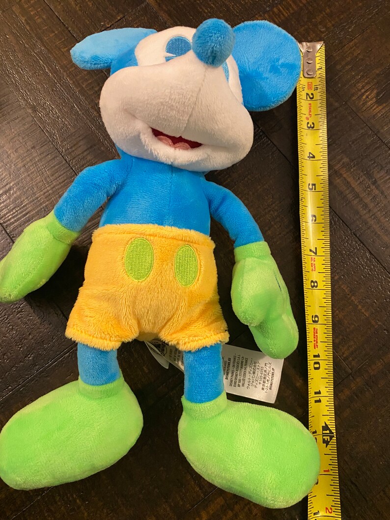 Op de afbeelding: Blauw en geel pluche Mickey Mouse speelgoed met groene schoenen en een label op de voet. Het speelgoed is ongeveer 28 cm hoog.