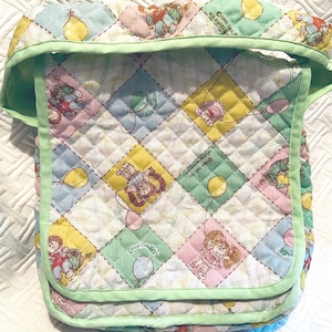 May include: A green quilted baby bib with a white background and a colorful pattern of Cabbage Patch Kids characters holding balloons. The bib has a green trim and the text "Cabbage Patch Kids" is printed on the bib.