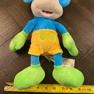 Op de afbeelding: Een blauw en groen pluche Mickey Mouse speelgoed met een gele broek. Het speelgoed is ongeveer 25 cm hoog en ligt op een houten oppervlak.