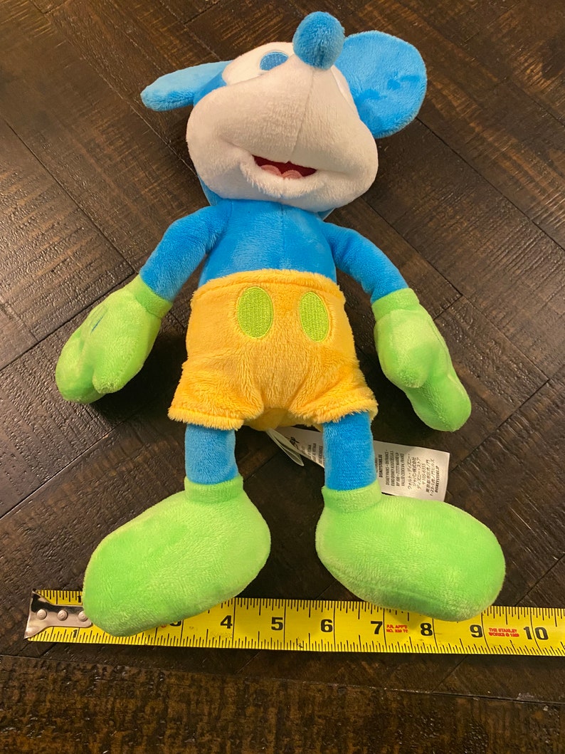 Op de afbeelding: Een blauw en groen pluche Mickey Mouse speelgoed met een gele broek. Het speelgoed is ongeveer 25 cm hoog en ligt op een houten oppervlak.