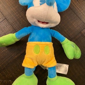 Op de afbeelding: Een blauw en groen pluche Mickey Mouse speelgoed dat een gele broek draagt. Het speelgoed heeft een wit gezicht met zwarte ogen en een grote glimlach.