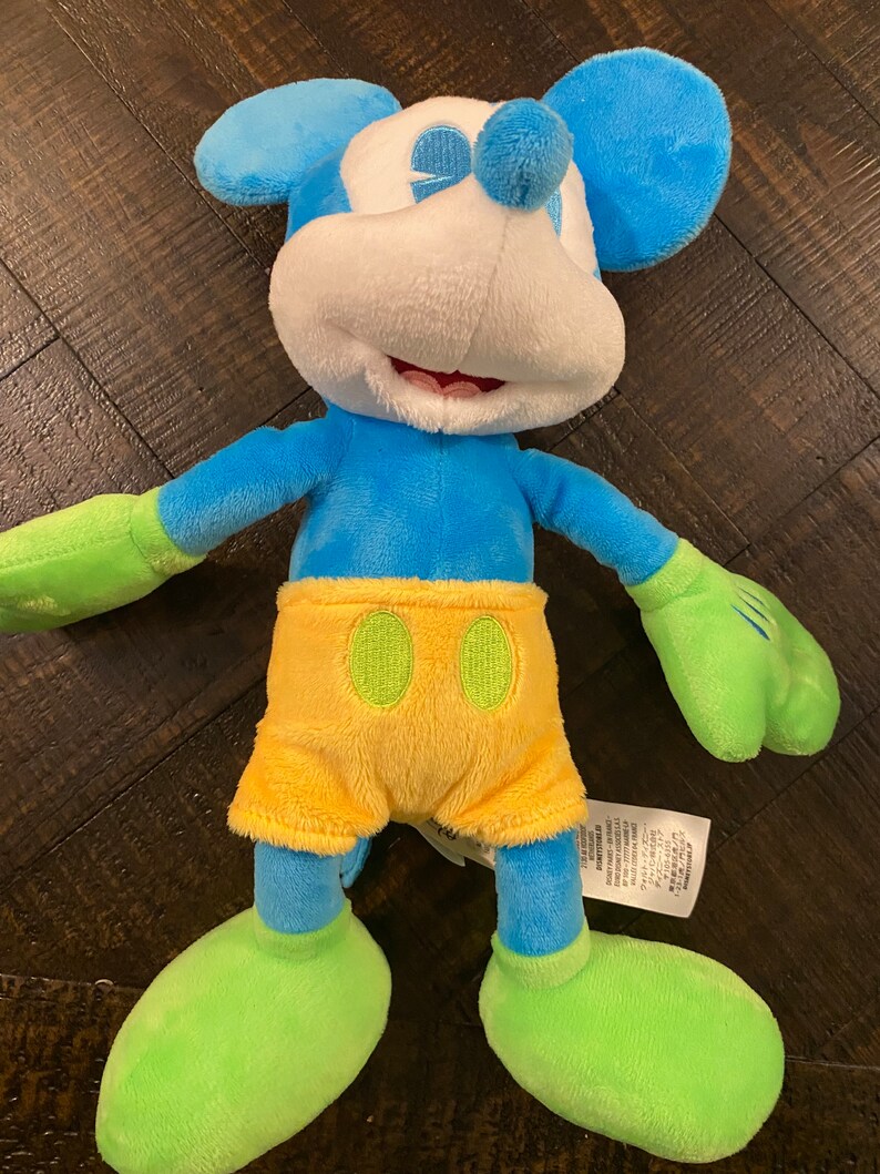 Op de afbeelding: Een blauw en groen pluche Mickey Mouse speelgoed dat een gele broek draagt. Het speelgoed heeft een wit gezicht met zwarte ogen en een grote glimlach.
