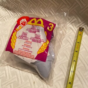 May include: A sealed plastic bag containing a Buzz Lightyear McDonald's Happy Meal toy. The packaging features the Disney Pixar Buzz Lightyear logo and the number 3. A yellow and black measuring tape is next to the bag.
