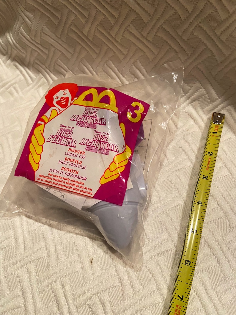 May include: A sealed plastic bag containing a Buzz Lightyear McDonald's Happy Meal toy. The packaging features the Disney Pixar Buzz Lightyear logo and the number 3. A yellow and black measuring tape is next to the bag.