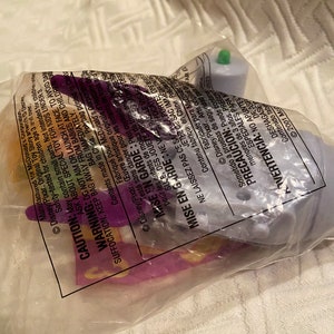 May include: A sealed plastic bag containing a Disney Pixar toy. The toy appears to be a small, light purple object with a green top. The bag has printed warnings and text in multiple languages.