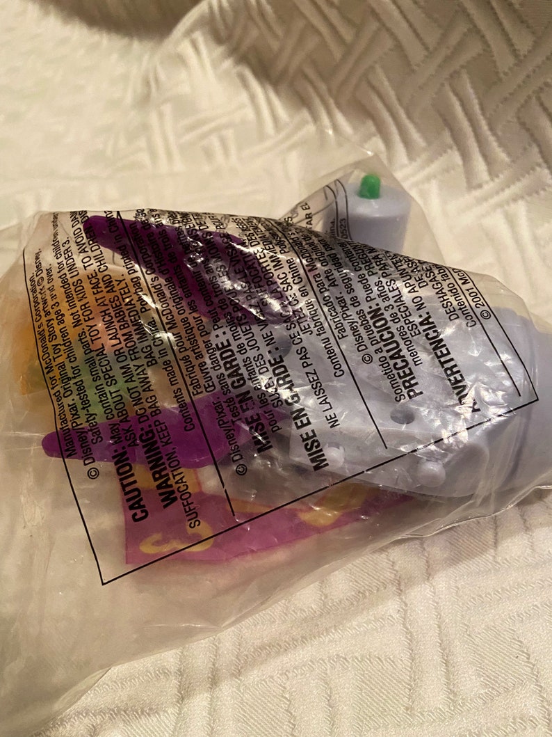 May include: A sealed plastic bag containing a Disney Pixar toy. The toy appears to be a small, light purple object with a green top. The bag has printed warnings and text in multiple languages.
