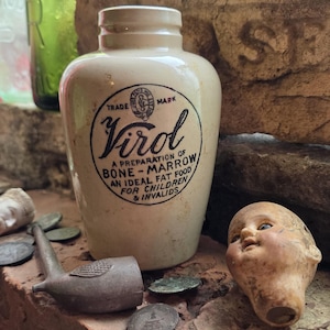 Puede incluir: Un tarro de cerámica de color crema con cuello estrecho y una etiqueta impresa en negro que dice: Trade Mark Virol, A Preparation of Bone-Marrow, An Ideal Fat Food for Children & Invalids. El tarro está junto a una cabeza de muñeca.