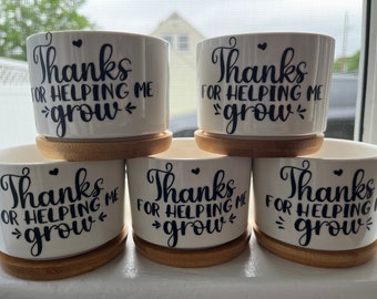 May include: Five white ceramic cylindrical planters, each resting on a circular bamboo saucer. The front of each planter features black script lettering that reads "Thanks FOR HELPING ME grow" with small heart and decorative accent symbols.