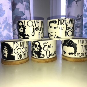 May include: Six white ceramic planters with black and white designs featuring characters and quotes from the TV show "Schitt's Creek". The quotes include "Love the journey,  "Are we a bad home?", "I'd kill for good coffee right now", "Ew, David", and "I have a son,  "Three, now four".