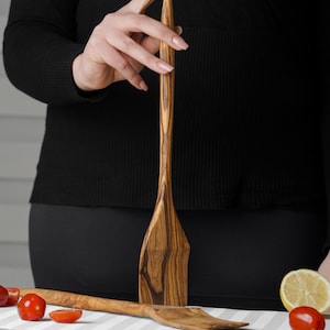 Könnte beinhalten: Zwei Holzlöffel für die Küche mit natürlichem Holzmaserungsmuster. Die Löffel sind aus Olivenholz und haben einen langen, schmalen Griff.
