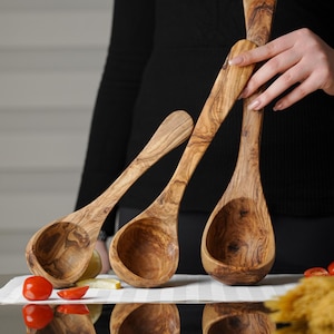 Puede incluir: Tres cucharones de madera de olivo de diferentes tama&ntilde;os. Los cucharones son marrones con un patr&oacute;n de veta de madera natural. Los cucharones est&aacute;n dispuestos en forma triangular con el cuchar&oacute;n m&aacute;s grande en la parte inferior.