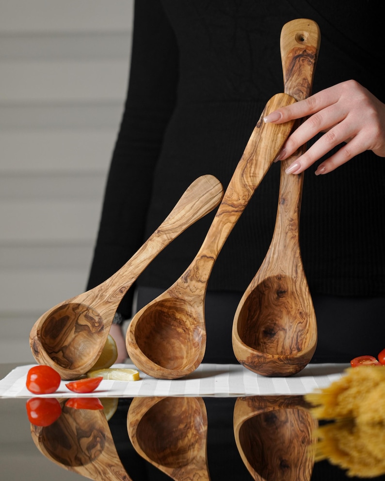 Puede incluir: Tres cucharones de madera de olivo de diferentes tama&ntilde;os. Los cucharones son marrones con un patr&oacute;n de veta de madera natural. Los cucharones est&aacute;n dispuestos en forma triangular con el cuchar&oacute;n m&aacute;s grande en la parte inferior.