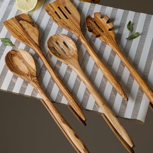 May include: A set of six wooden kitchen utensils with natural wood grain patterns. The utensils include a slotted spoon, a spatula, a pasta server, and three spoons. The utensils are arranged on a white and gray striped surface.