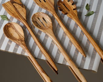 May include: A set of six wooden kitchen utensils with natural wood grain patterns. The utensils include a slotted spoon, a spatula, a pasta server, and three spoons. The utensils are arranged on a white and gray striped surface.