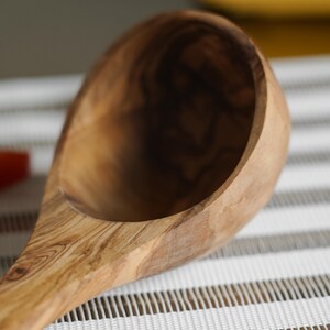 Puede incluir: Un cuchar&oacute;n de madera de olivo con un patr&oacute;n de veta de madera natural. El cuchar&oacute;n est&aacute; apoyado sobre una superficie rayada blanca y marr&oacute;n.