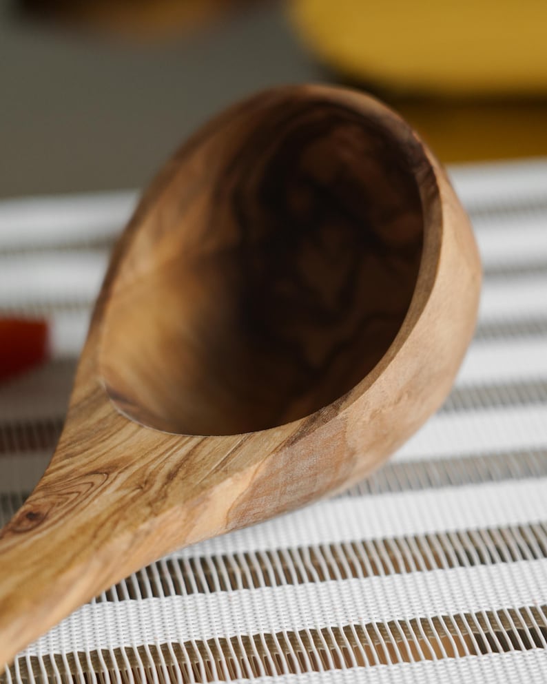 Puede incluir: Un cuchar&oacute;n de madera de olivo con un patr&oacute;n de veta de madera natural. El cuchar&oacute;n est&aacute; apoyado sobre una superficie rayada blanca y marr&oacute;n.
