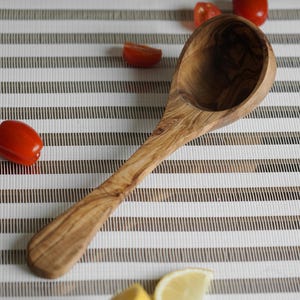 Puede incluir: Un cuchar&oacute;n de madera de olivo con un acabado de grano de madera natural. El cuchar&oacute;n est&aacute; sobre una superficie rayada blanca y marr&oacute;n.
