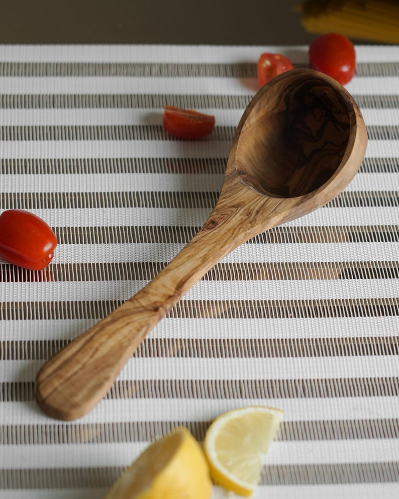 Puede incluir: Un cuchar&oacute;n de madera de olivo con un acabado de grano de madera natural. El cuchar&oacute;n est&aacute; sobre una superficie rayada blanca y marr&oacute;n.