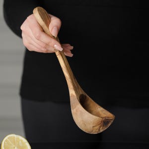 Puede incluir: Un cuchar&oacute;n de madera de olivo con un mango largo. El cuchar&oacute;n se sostiene en la mano de una persona.