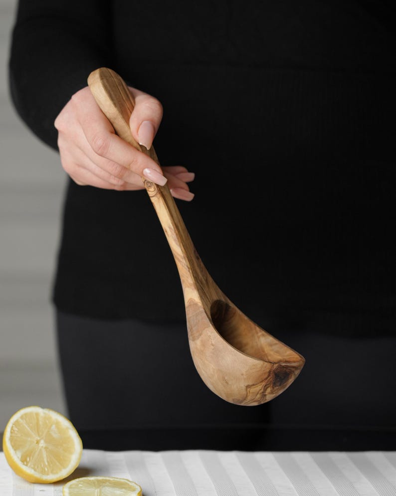 Puede incluir: Un cuchar&oacute;n de madera de olivo con un mango largo. El cuchar&oacute;n se sostiene en la mano de una persona.